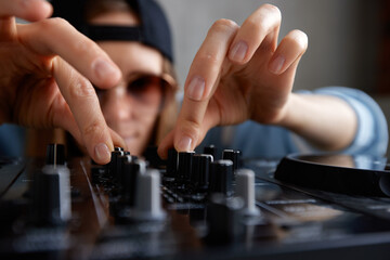 A young pretty long-haired DJ girl in a blue sweater and black baseball cap poses with a black DJ mixing console and mixes music tracks. Close-up studio shot, gray background.