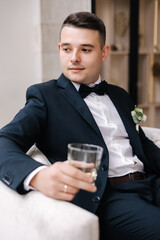 Handsome groom in dark blue suit sitting on chair with glass of whiskey. Groom's morning. Wedding day