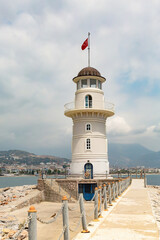 Fototapeta na wymiar Lighthouse in the port of Alanya. White lighthouse in seaport of Alanya with concrete path and stones, scenic clouds as background. Vertical shot. Turkey (Turkiye)