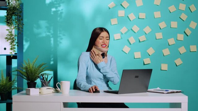Injured woman wearing cervical collar cast struggling while working on laptop in office. Wounded employee with neck brace being in pain at colourful workplace desk over blue studio background