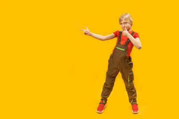 Teenage boy in overalls poses in studio on yellow background and points fingers to the side. Copy space. Space for text.