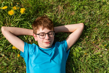 Boy wears glasses and lies on green lawn with his hands behind his head. Happy child lying on green...