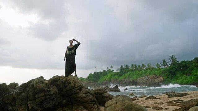 Godlike gender fluid person of color in luxury dress, jewelry stands on cliff peak at scenic ocean. Pride extravagant fashion model in epic outfit posing like goddess in tropics. Olymp concept.