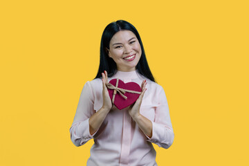 Young cheerful korean woman holding heart shape gift box in both hands. Isolated on yellow background.