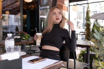 Brooding blonde woman sits in coffee shop with cup of cappuccino coffee latte. Attractive girl alone in cafe