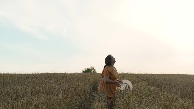Middle aged European free woman throws up her hat over ripe grain field. Lady plus size in orange dress enjoys her vacation, relaxes in nature. Concept of self acceptance, care in mental health.