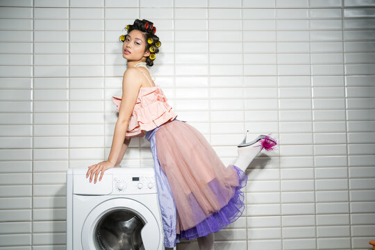 asian young woman with hair curlers standing in pink ruffled top, pearl necklace, tulle skirt and high heel shoes with feather near modern washing machine near white tiles in laundry room