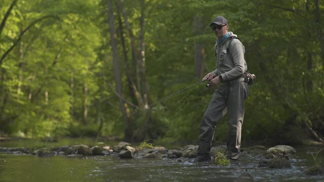 Fisherman catching brown trout on spinning tackle standing in river.