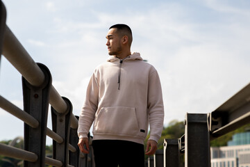 A young man in a sweatshirt stands on a narrow bridge against a cloudy sky. An Asian guy in a white sweatshirt stands on a bridge and looks away.