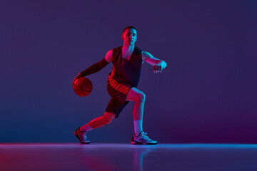 Fototapeta na wymiar Young sportive man, basketball player in uniform during game, dribbling ball against purple studio background in neon light. Concept of professional sport, hobby, healthy lifestyle, action and motion