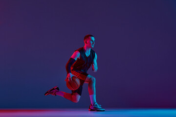 Concentrated young man, basketball player during game, training, dribbling ball against purple...