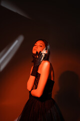 attractive asian woman with wet hairstyle posing in strapless dress with tulle skirt and black gloves with rings while standing on dark orange background with red lighting, model, looking at camera