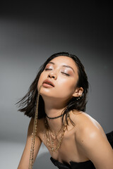 stylish asian model with short brunette hair holding golden jewelry in mouth, posing in strapless dress on grey background, everyday makeup, wet hairstyle, young woman, closed eyes, portrait