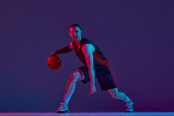 Fototapeta na wymiar Young muscular man, basketball player in motion, dribbling ball against purple studio background in neon light. Winner. Concept of professional sport, hobby, healthy lifestyle, action and motion