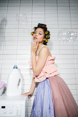 young asian woman with hair curlers standing in ruffled top, pearl necklace and tulle skirt near modern washing machine with detergent bottle in laundry room, housewife, soap bubbles