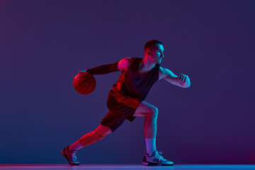Young athletic man, basketball player in motion with ball against purple studio background in neon light. Champion. Concept of professional sport, hobby, healthy lifestyle, action and motion