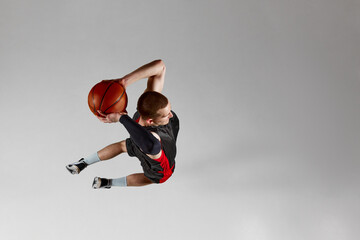 Motivated, concentrated young man, basketball player throwing all in a jump against grey studio background. Aerial view. Slam dunk. Concept of professional sport, competition, action and motion © master1305