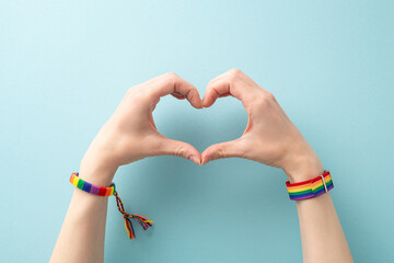 LGBT support idea. First-person photo of female hands donning symbolic rainbow bracelets creating heart gesture on pastel blue background with ample space for text or ad