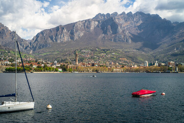 Buildings of the city of Lecco on the shores of Lake Como surrounded by mountains in the background. Sailboats on the waters of the lake and the waterfront.