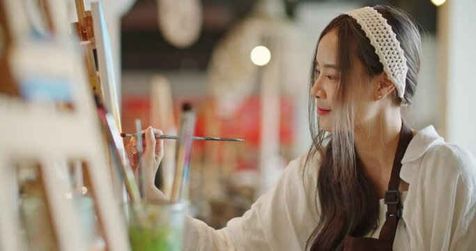 Beautiful asian stylish female artist painting picture in art studio. Young woman painter draws with paint brush and oil colors creating artwork sunset landscape on canvas. imagination and creativity.