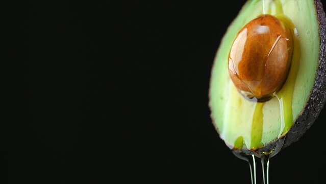 Avocado oil pouring, dripping essential oil on cut avocado isolated on black background, slow motion. Fresh cut avocado with oil stream. Concept of healthy fruit for eating or cosmetics. 