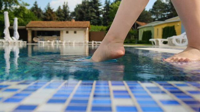 A young girl is testing the temperature of the water in the pool with her foot. Close-up.