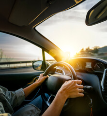 Female hands on the steering wheel of a car. Woman driving a car at sunset on the seashore. Auto...