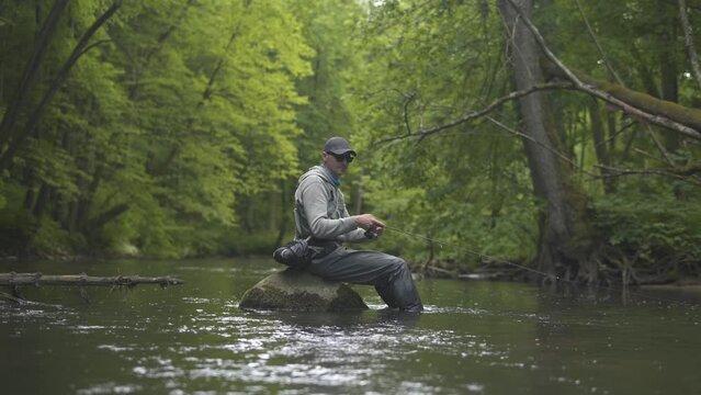 Fisherman catching brown trout on spinning tackle sitting on rock in river. 