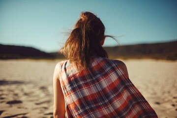 Brown haired girl covered with a cloth plaid cape standing peacefully at the beach on the sand and looking into the distance, generative AI