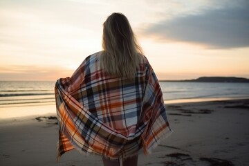Blonde haired girl covered with a cloth plaid blanket standing peacefully at the beach and looking into the ocean , generative AI