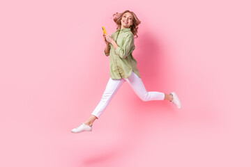 Full length photo of excited funky lady wear green shirt typing messages modern gadget jumping high isolated pink color background