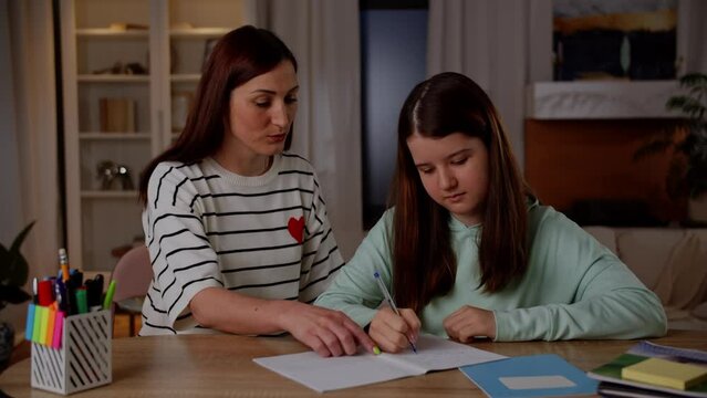 A brunette woman in a striped sweater helps her daughter with her homework. Mom praises and hugs her daughter who did her homework.