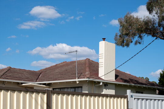 An old brick Australian residential house with roof tiles and a chimney behind metal garden fences. Concept of home ownership,  household privacy, and real estate or housing market.