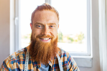 handsome redhaired man with long beard sews at a sewing machine at home studio