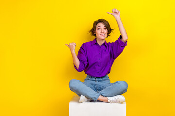 Full length photo of pretty nervous person sit podium biting lips look indicate fingers empty space isolated on yellow color background