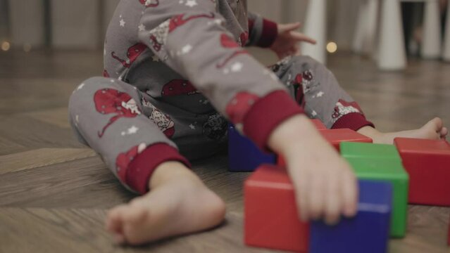 kid toddler sits floor gray pajamas plays multi-colored plastic cubes builds tower destroys with feet and hands, child learns break and build. problem upbringing, disobedience, crisis children concept