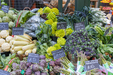 Fresh vegetables on a stand at a food market in the UK including, Romanesco, artichoke, corn, cauliflower, and asparagus. No people.
