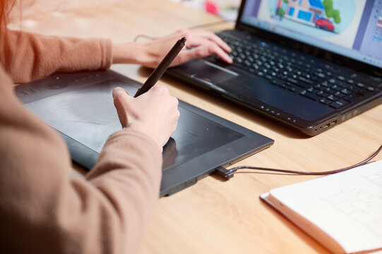 Closeup look at a female illustrator working on her laptop and using a graphic tablet to create a fresh illustration for a company's rebranding. She is engaged in remote work, online work, deadline.