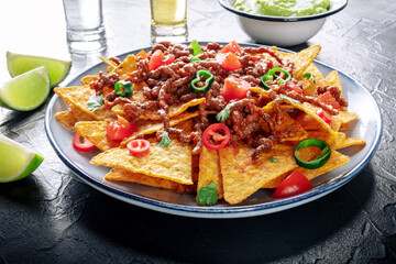Nachos, Mexican food, tortilla chips with beef and fresh vegetables, with tequila, guacamole, and limes, on a black slate background