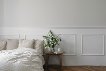 Cup of coffee, tea on wooden night stand. Green bouquet of white viburnum, fern and solomons seal flowers. Bedroom view. Beige pillows, linen blanket in bed. Elegant moulding. Empty wall mockup. - obrazy, fototapety, plakaty