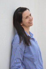 Fototapeta premium A young woman with dark hair in a blue collared shirt, stands against a white wall, smiles and looks into the distance.