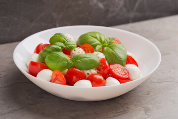 Caprese salad with tomato, mozzarella, basil and olive oil on white plate on dark table. Isometric style. Mediterranean cuisine.