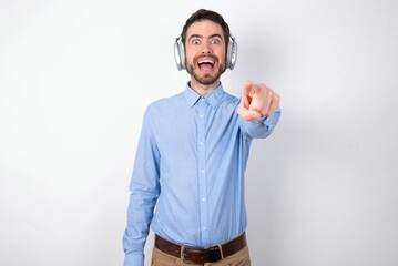 Excited positive businessman wearing blue t-shirt with headphones over white background points index finger directly at you, sees something very funny. Wow, amazing