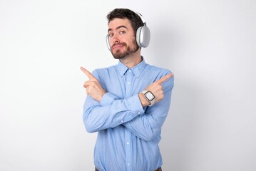 businessman wearing blue t-shirt with headphones over white background crosses arms and points at different sides hesitates between two items or variants. Needs help with decision