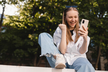 Smiling young girl sitting on ground browsing internet with cellphone gesturing like winner....