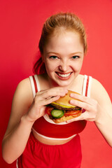 Beautiful hungry woman, girl holding and eating a juicy burger with desire over red studio background. Usable for food advertising. Close up