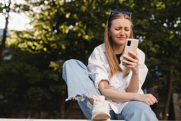 Pretentious blonde woman hold mobile phone and gloomy laughing with disgusting. Girl with a grin, sarcasm sarcastically look. Woman sitting in city park, wear white shirt and blue jeans. - obrazy, fototapety, plakaty