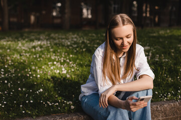 Young stylish blonde woman writes message on smartphone sitting on green city park, hipster tourist traveling in town and planning trip using mobile phone. Look smiling when read message.