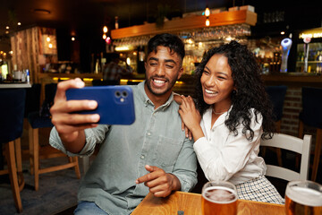 Young multiracial couple in casual clothing smiling and taking selfie with mobile phone at bar