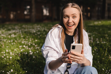 Portrait of astonished young blonde girl sitting on ground hold phone open mouth wear white shirt, black top. Wow, omg, good news, emotions walk outdoors in the park. Win in lottery. Unbelievable!